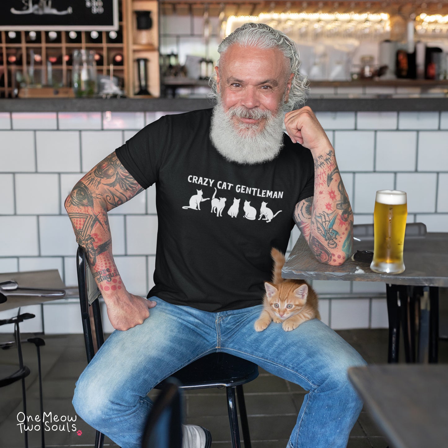 A man with a white beard and tattoos sits in a bar setting wearing a black graphic t-shirt that reads 'CRAZY CAT GENTLEMAN' with cat silhouettes. An orange tabby kitten rests on his lap, and a glass of beer is on the table next to him.