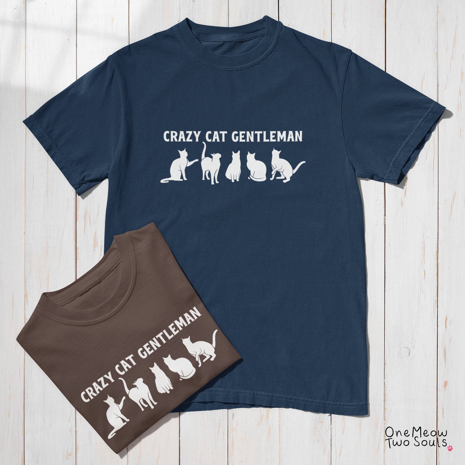 A navy blue short-sleeve Comfort colors 1717 t-shirt and a brown short-sleeve t-shirt, both featuring a white graphic print that reads 'CRAZY CAT GENTLEMAN' above a row of cat silhouettes. The shirts are arranged on a white wood plank background.