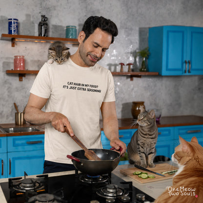 A man in a kitchen wearing a white graphic t-shirt that reads 'CAT HAIR IN MY FOOD? IT'S JUST EXTRA SEASONING'. Cats are present around him, including one on his shoulder, one sitting on the counter, and another in the foreground. One Meow Two Souls brand.