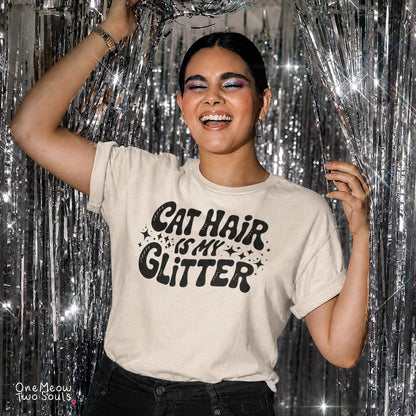 A woman wearing a beige graphic t-shirt with the text 'CAT HAIR IS MY GLITTER' in black, posing in front of a silver tinsel curtain. The brand name 'One Meow Two Souls' is visible in the bottom-left corner.