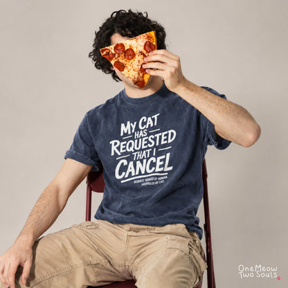 A man wearing a navy blue graphic t-shirt with the text 'MY CAT HAS REQUESTED THAT I CANCEL' sits on a red chair holding a slice of pepperoni pizza to his face. The background is a solid light gray studio wall. One Meow Two Souls brand.