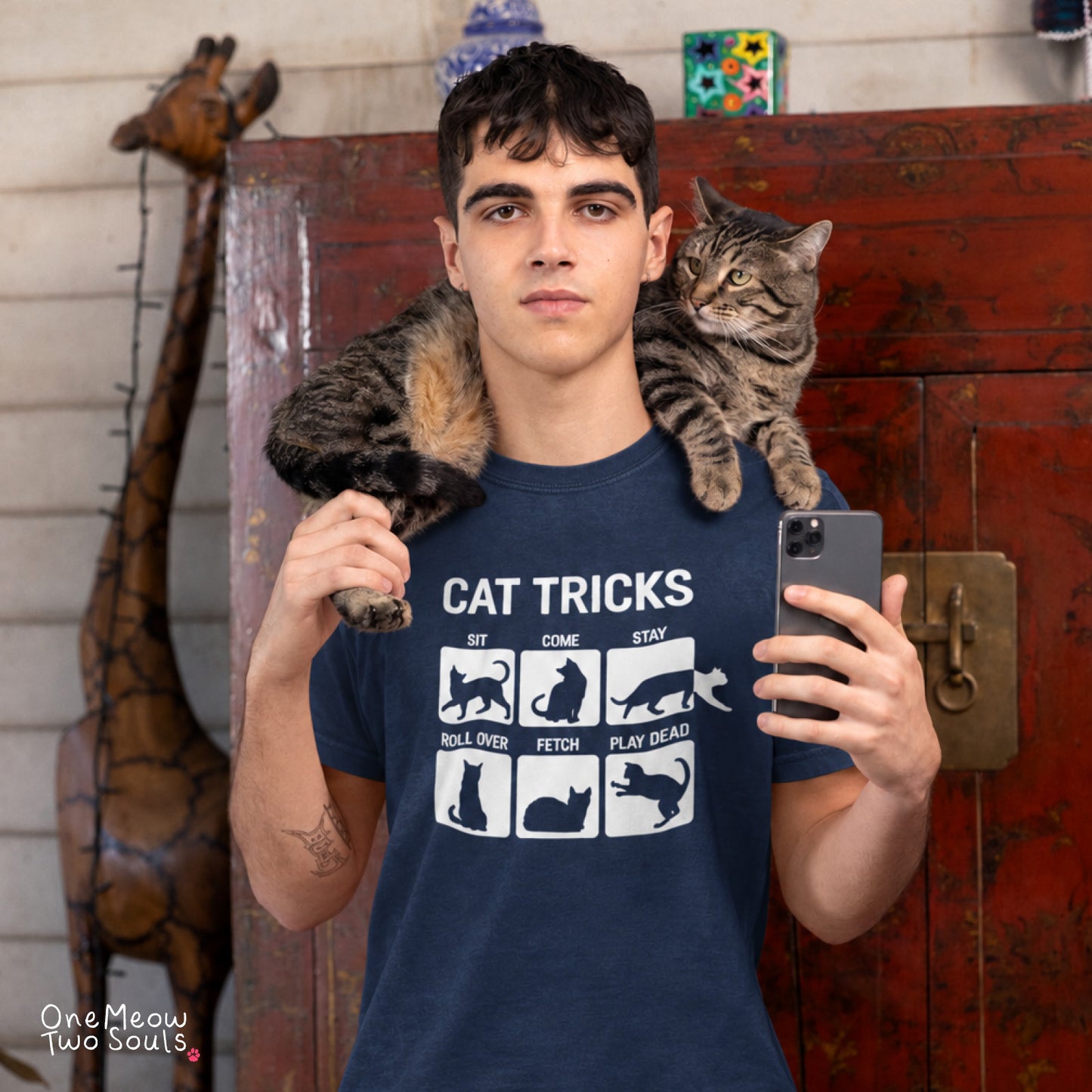 A man wearing a navy blue graphic t-shirt with a white cat print that reads 'CAT TRICKS' and showing a tabby cat on his shoulders, taking a mirror selfie in front of a rustic red wooden cabinet. One Meow Two Souls brand.