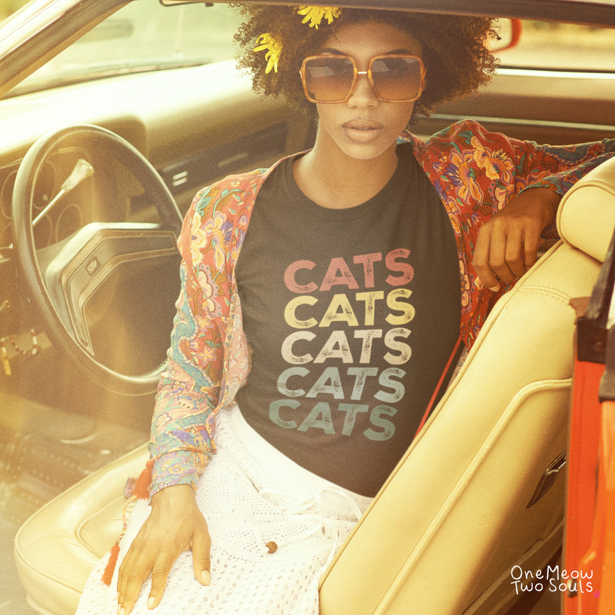 A woman sitting inside a vintage car wearing a black graphic t-shirt with the word 'CATS' repeated five times in colorful gradient letters, styled with a multicolor paisley cardigan, white pants, and sunglasses.
Black colored Comfort Colors 1717 cotton T-shirt, featuring a retro "CATS" cool cat design illustration. One Meow Two Souls brand.