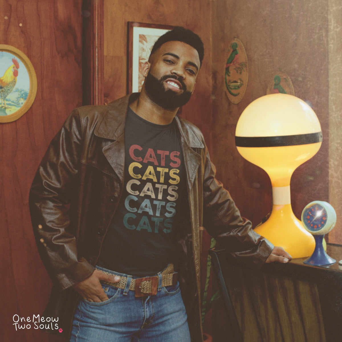 A man wearing a brown leather jacket and a black graphic t-shirt with the word 'CATS' repeated in colorful letters, leaning against a table in a retro-themed room with a yellow lamp.
Black colored Comfort Colors 1717 cotton T-shirt, featuring a retro "CATS" cool cat design illustration. One Meow Two Souls brand.