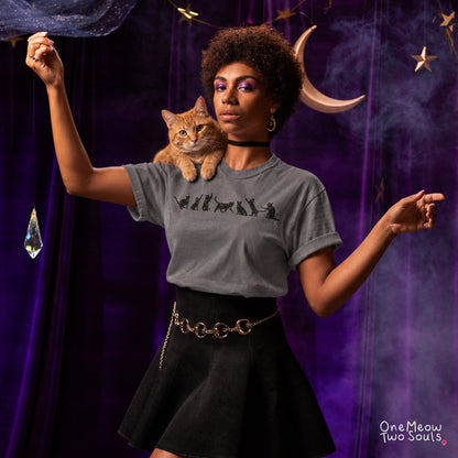 A woman with curly hair wearing a gray graphic t-shirt and a black skirt, with an orange tabby cat on her shoulder. She is posing in front of a dark purple curtain decorated with a crescent moon, stars, and a hanging crystal. One Meow Two Souls brand.
