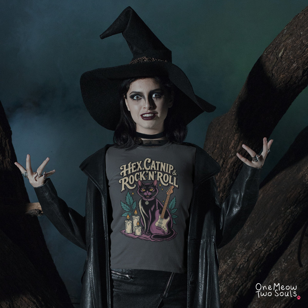 A person wearing a witch hat, black leather-like costume jacket, and a dark graphic T-shirt stands between large tree trunks in a moody outdoor setting. The shirt graphic reads 'Hex Catnip Rock N Roll' with a cat, candles, and a guitar illustration. One Meow Twou Souls brand.