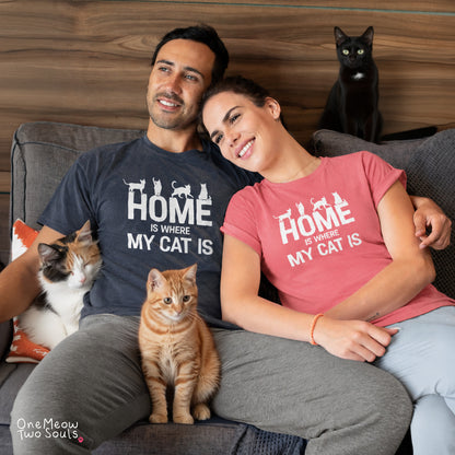 A man and woman sitting on a couch wearing matching graphic t-shirts that read 'HOME IS WHERE MY CAT IS' with cat silhouettes. A calico cat rests on the man's lap, an orange tabby sits in front of them, and a black cat sits on the couch behind them. One Meow Two Souls brand.