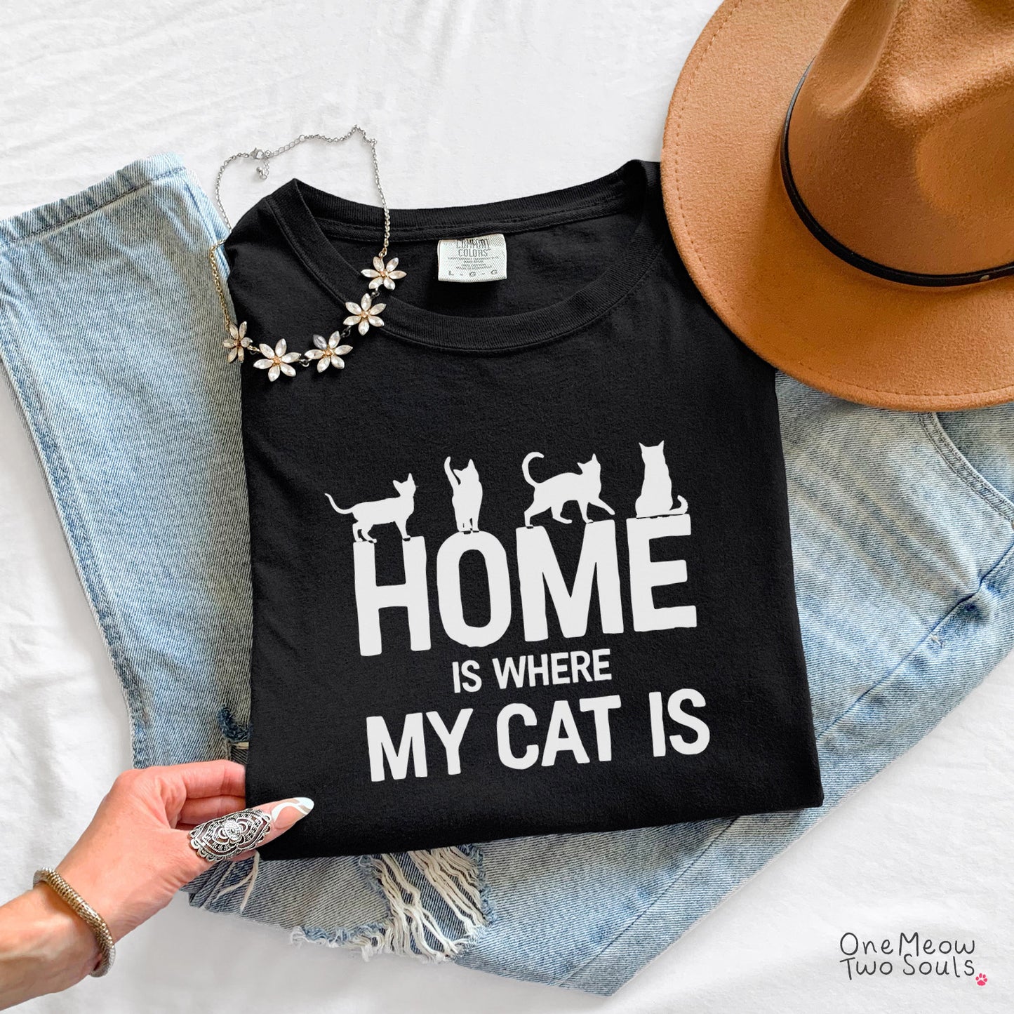 A black graphic T-shirt laid flat on top of light blue jeans, with a tan hat, white flowers, and a hand visible holding the shirt. The shirt reads “HOME IS WHERE MY CAT IS” in white text with cat silhouettes above the words. One Meow Two Souls brand.