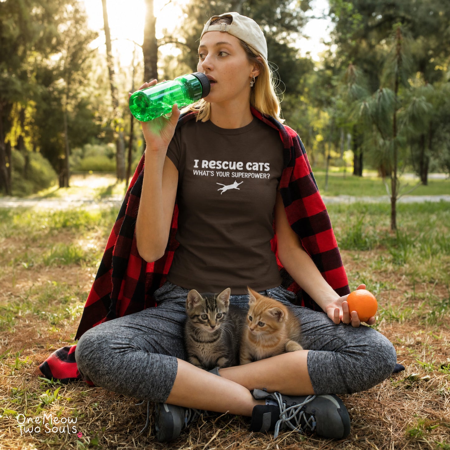 A woman sitting outdoors in a park with two kittens on her lap, wearing a brown graphic t-shirt that reads 'I Rescue cats WHAT'S YOUR SUPERPOWER?'. She has a red and black buffalo plaid shirt draped over her shoulders and is drinking from a green bottle. One Meow Two Souls brand.