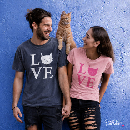 A man and a woman wearing matching graphic t-shirts with 'LOVE' and a cat face, standing against a blue textured wall with an orange tabby cat on their shoulders. The man's shirt is dark gray with white print, and the woman's shirt is pink with a pink print. One Meow Two Souls brand.