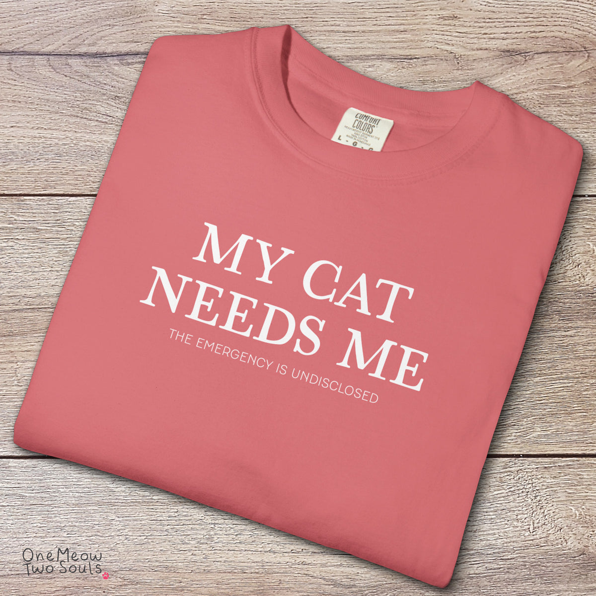 Watermelon colored Comfort Colors 1717 cotton T-shirt, featuring a "My Cat Needs Me. The Emergency is Undisclosed" funny cat design illustration. One Meow Two Souls brand.