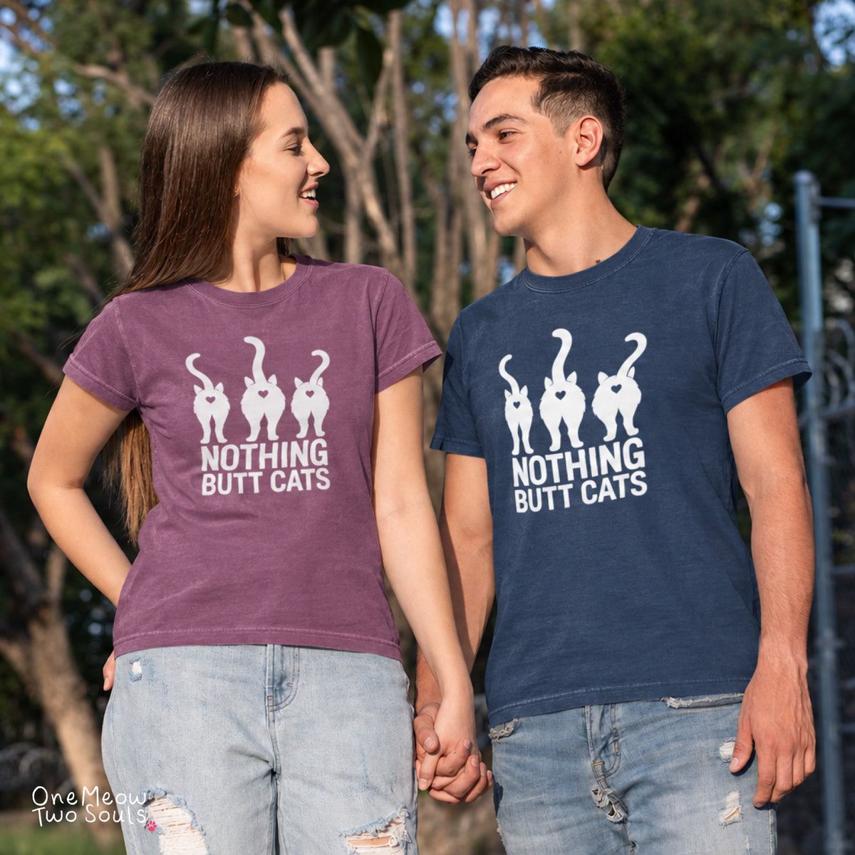 A man and a woman standing outdoors and holding hands, both wearing matching graphic t-shirts with the white text 'NOTHING BUT CATS' and four cat silhouettes. The woman's shirt is a heathered purple, and the man's shirt is navy blue. One Meow Two Sould brand.