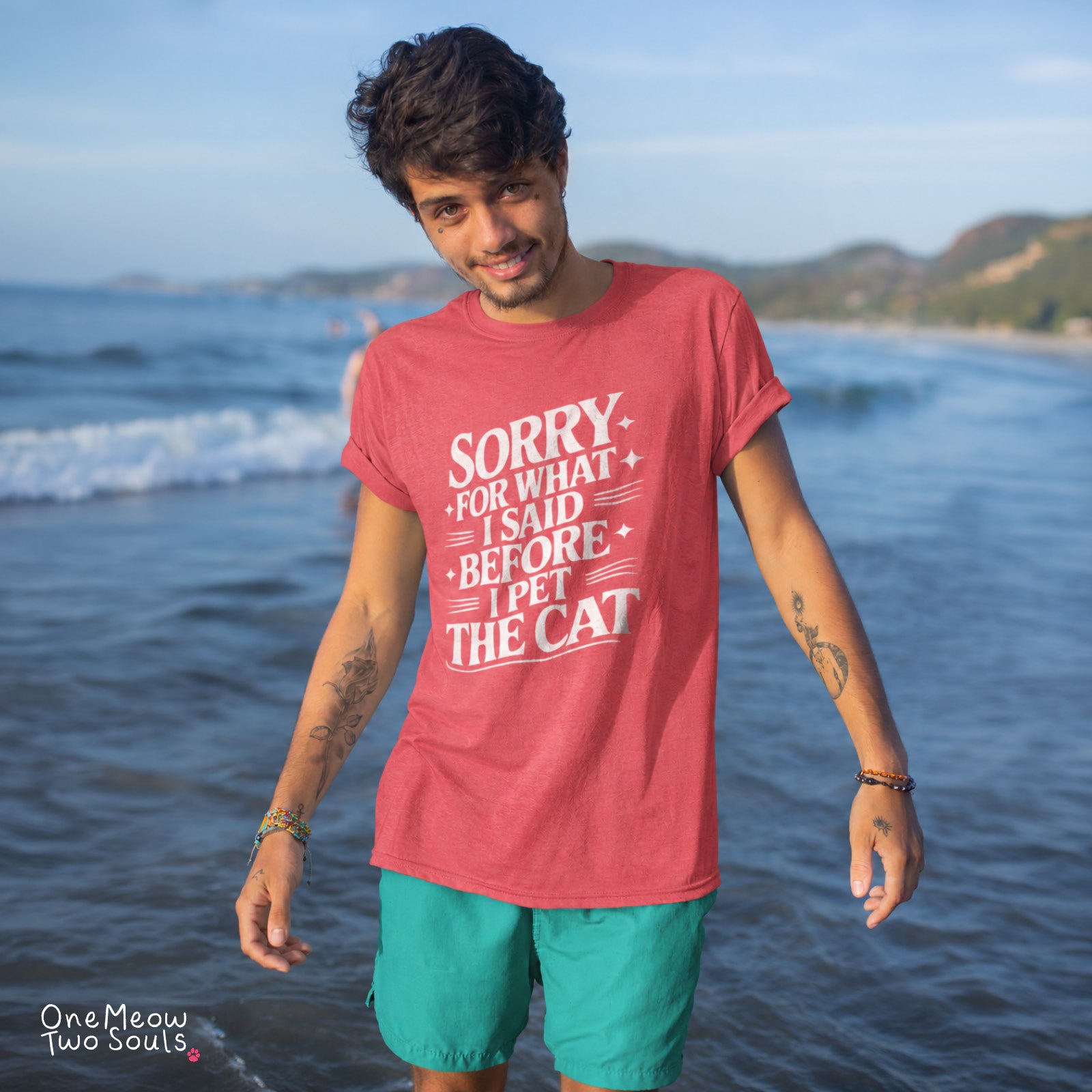 A man standing on a beach wearing a coral red graphic t-shirt with white text that reads 'SORRY FOR WHAT I SAID BEFORE I PET THE CAT' and teal shorts. The brand name 'One Meow Two Souls' is visible in the bottom left corner. One Meow Two Souls brand.