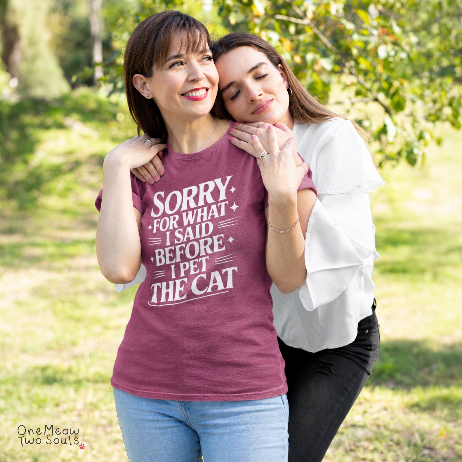 Two women hugging outdoors in a grassy park setting. The woman on the left wears a heathered maroon t-shirt with a white graphic text that reads 'SORRY FOR WHAT I SAID BEFORE I PET THE CAT'. One Meow Two Souls brand.