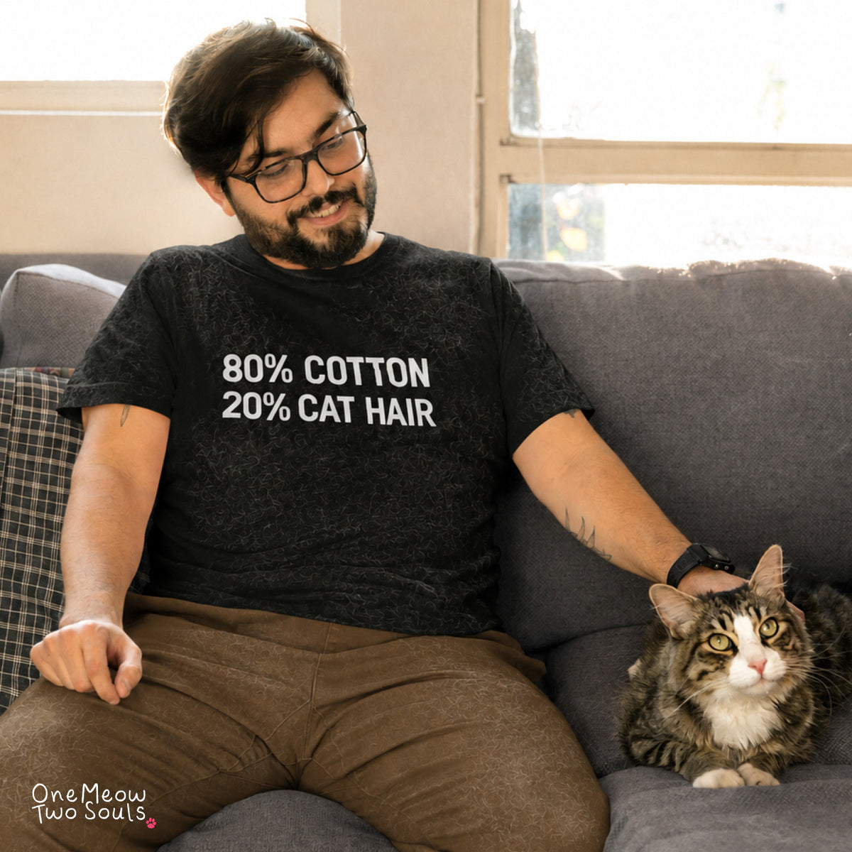 A man wearing a dark gray graphic t-shirt that reads '80% COTTON 20% CAT HAIR' sits on a gray couch with a tabby cat next to him. 
Black Color Comfort Colors 1717 t-shirt with a "80% cotton 20% cat hair" design. One Meow Two Souls brand.