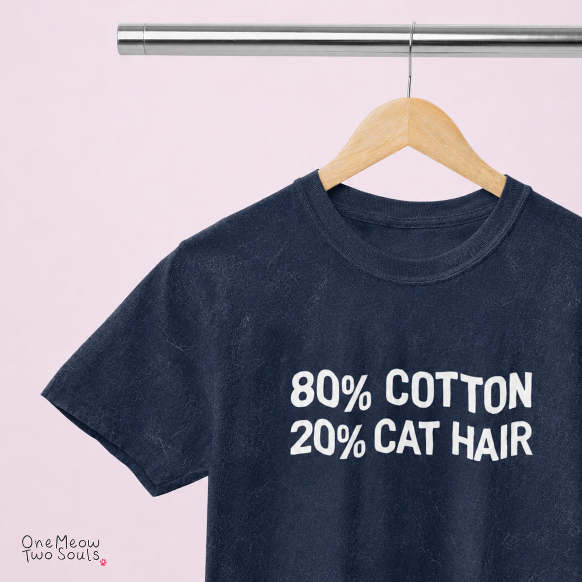A dark navy blue t-shirt hanging on a wooden hanger from a metal clothing rack, against a solid light pink background. The shirt features the text '80% COTTON 20% CAT HAIR' printed in white on the front. 
Navy Color Comfort Colors 1717 t-shirt with a "80% cotton 20% cat hair" design. One Meow Two Souls brand.
