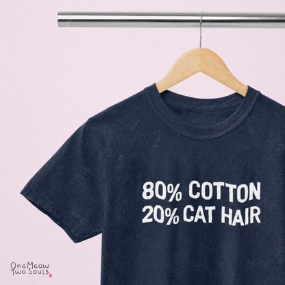 A dark navy blue t-shirt hanging on a wooden hanger from a metal clothing rack, against a solid light pink background. The shirt features the text '80% COTTON 20% CAT HAIR' printed in white on the front. 
Navy Color Comfort Colors 1717 t-shirt with a "80% cotton 20% cat hair" design. One Meow Two Souls brand.