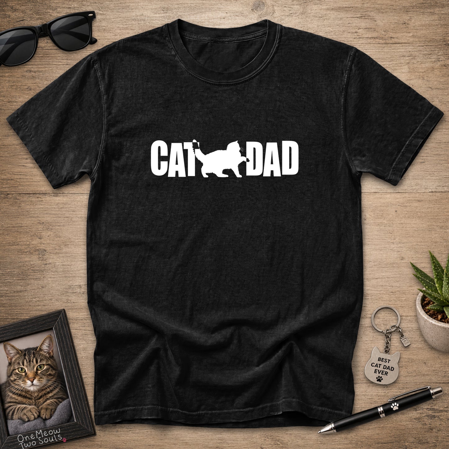 A black short-sleeve t-shirt laid flat on a wooden surface, featuring a black graphic print that reads 'CAT DAD' with a cat silhouette. The shirt is styled with sunglasses, a framed cat portrait, a small potted succulent, a keychain, and a pen.