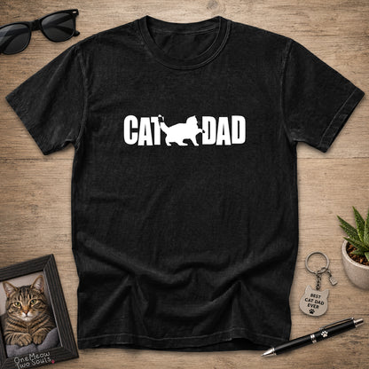 A black short-sleeve t-shirt laid flat on a wooden surface, featuring a black graphic print that reads 'CAT DAD' with a cat silhouette. The shirt is styled with sunglasses, a framed cat portrait, a small potted succulent, a keychain, and a pen.