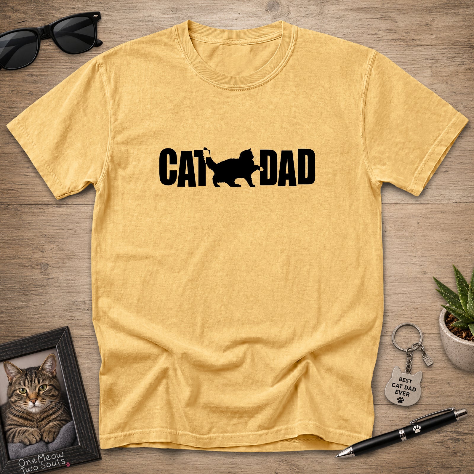 A mustard t-shirt with a black 'CAT DAD' graphic featuring a cat silhouette, displayed in a flat-lay arrangement on a wooden surface with sunglasses, a framed cat photo, a pen, and a small plant.