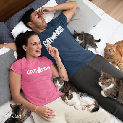 A man and a woman lying on a bed surrounded by several cats, wearing matching graphic T-shirts. The woman wears a bright pink T-shirt with the text 'CAT MOM' and a cat silhouette, while the man wears a navy blue T-shirt with the text 'CAT DAD' and a cat silhouette.