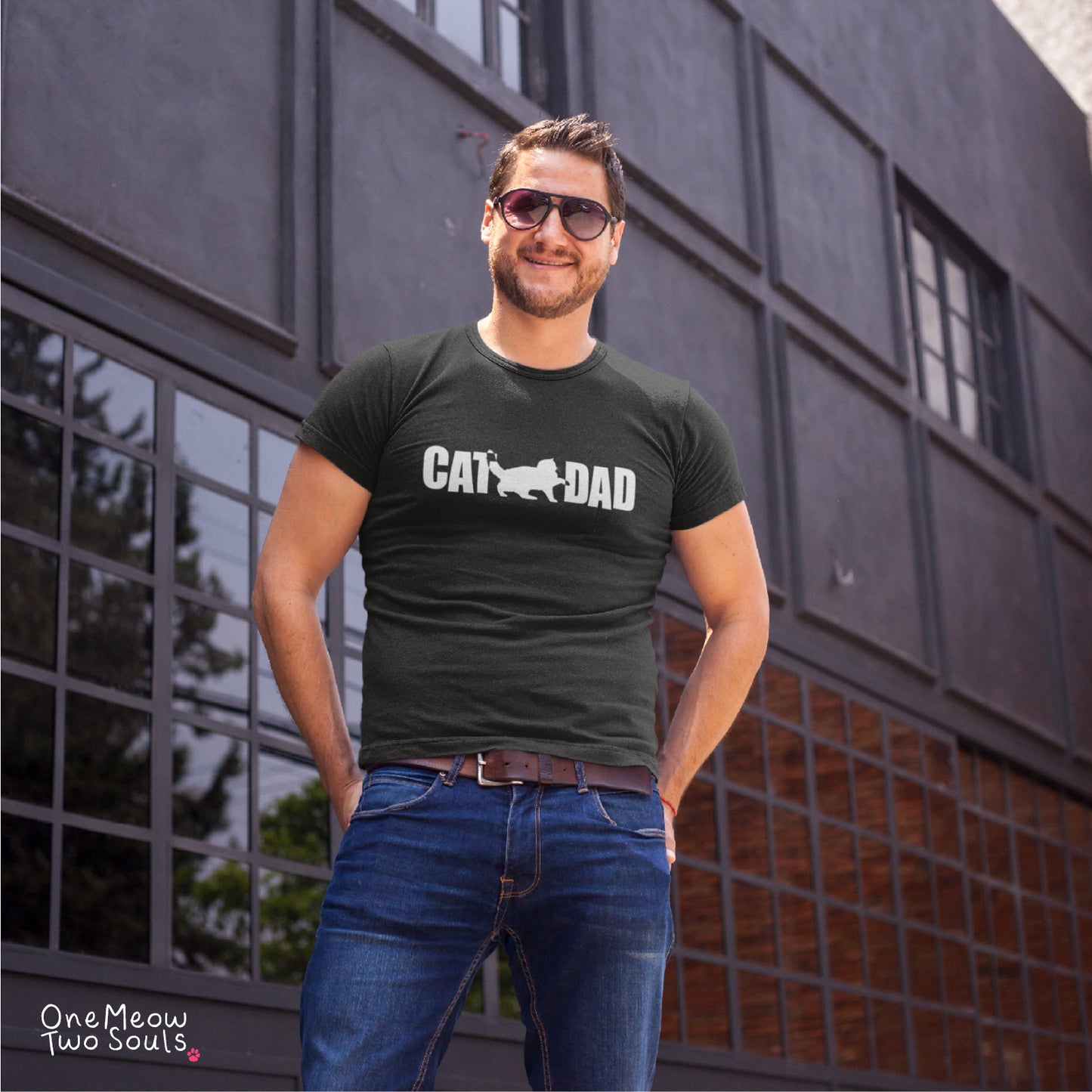 A man wearing a dark gray graphic t-shirt with 'CAT DAD' printed in white across the chest, paired with blue jeans and sunglasses, standing in front of a modern gray building.