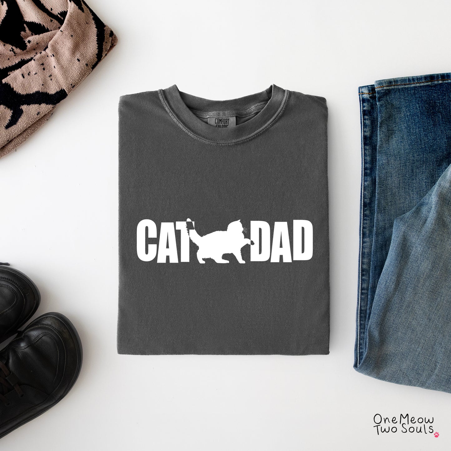 A folded dark gray t-shirt with a white graphic reading 'CAT DAD' and a cat silhouette, styled in a flat lay with jeans, black shoes, and a patterned scarf on a white background.