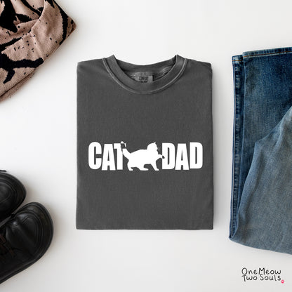 A folded dark gray t-shirt with a white graphic reading 'CAT DAD' and a cat silhouette, styled in a flat lay with jeans, black shoes, and a patterned scarf on a white background.