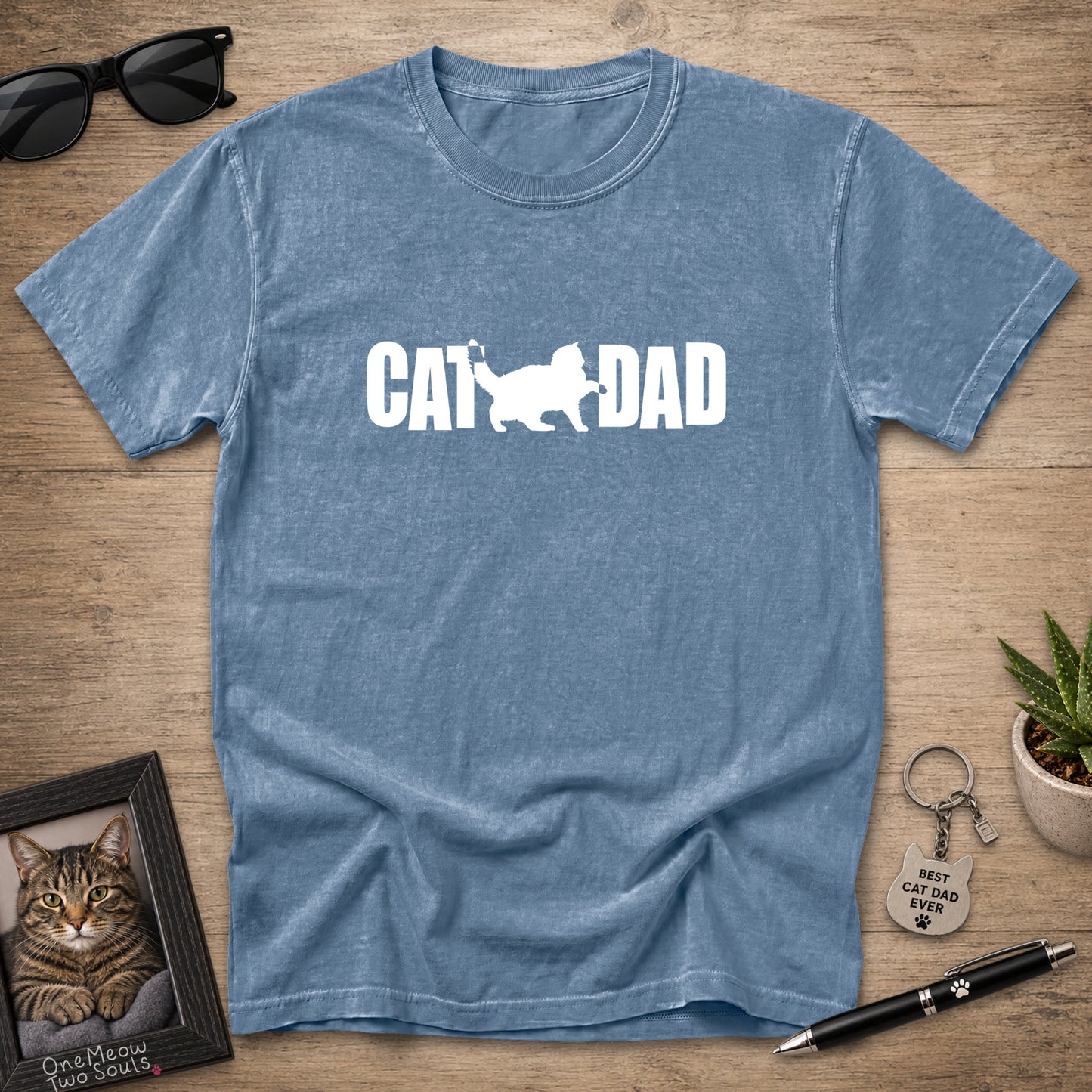 A heathered blue short-sleeve t-shirt laid flat on a wooden surface, featuring a white graphic that reads 'CAT DAD' with a cat silhouette. Sunglasses, a framed cat portrait, a small plant, a keychain, and a pen are arranged around it as props.