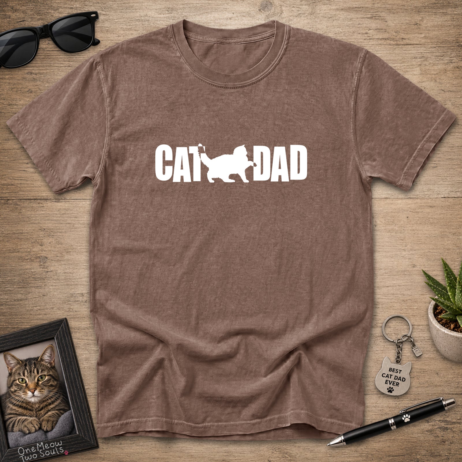 A brown short-sleeve t-shirt laid flat on a wooden surface, featuring a white graphic that reads 'CAT DAD' with a cat silhouette. Sunglasses, a framed cat portrait, a small plant, and a pen are placed around it as props.