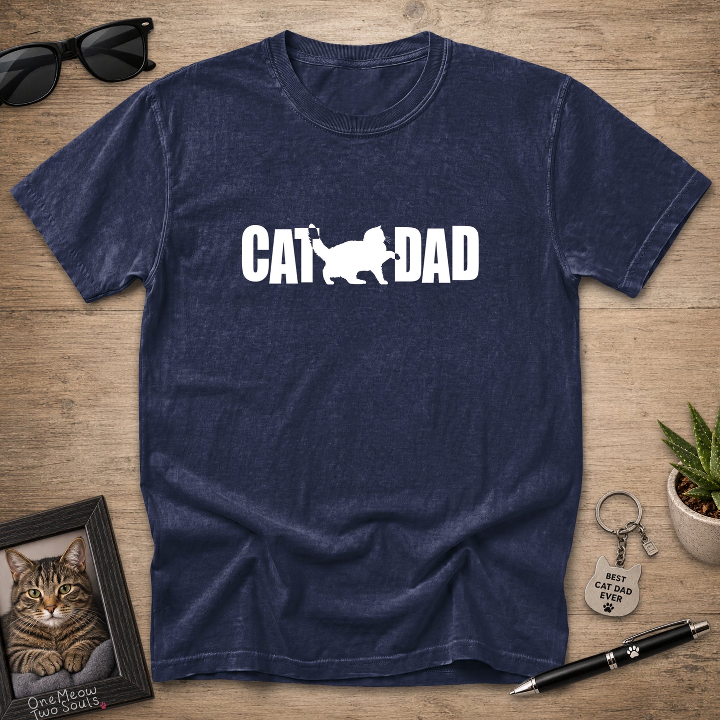 A navy blue heathered t-shirt with a white graphic reading 'CAT DAD' and a cat silhouette, laid flat on a wooden surface with sunglasses, a framed cat portrait, a small plant, and a pen around it.