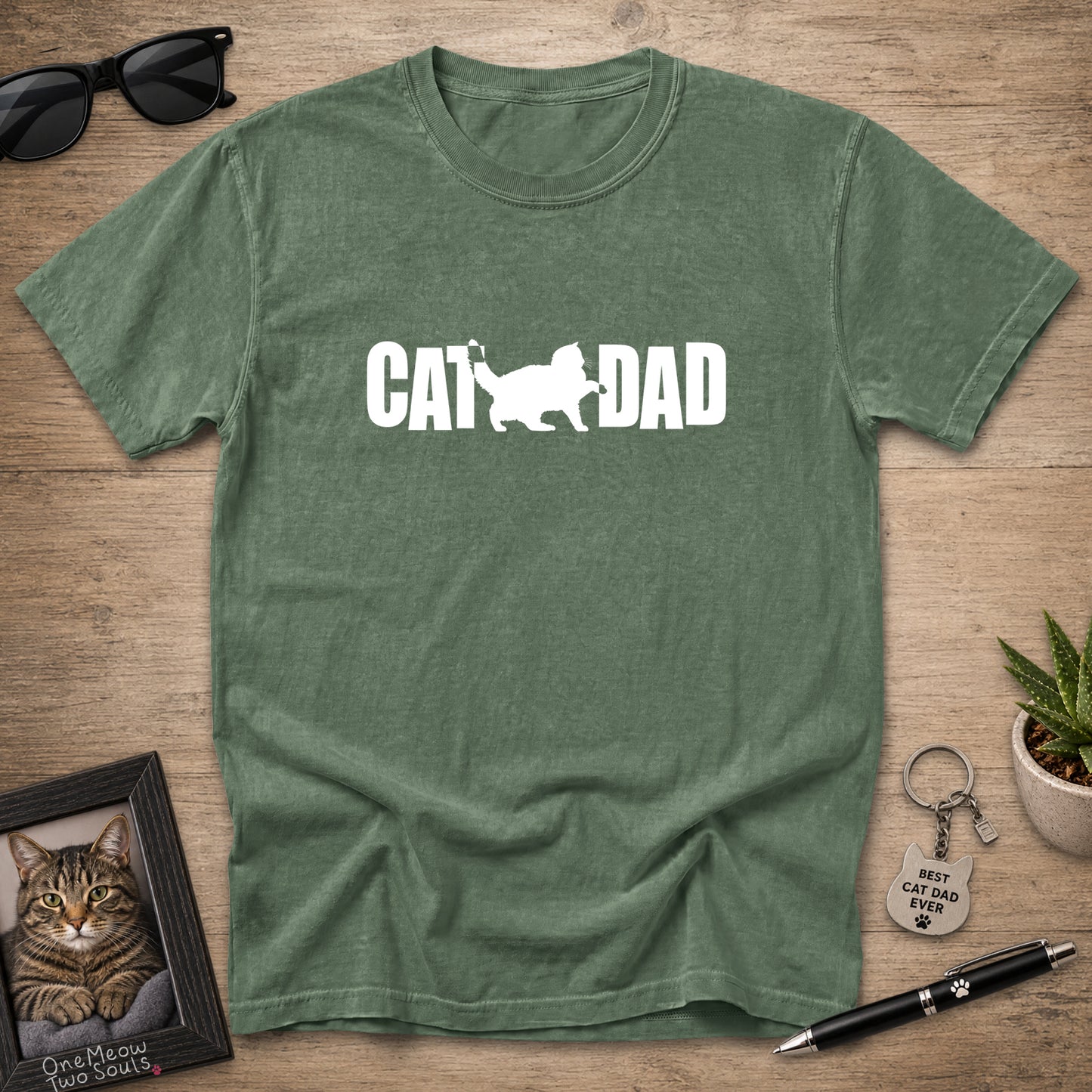 A heather green crewneck t-shirt with a white graphic reading 'CAT DAD' and a cat silhouette, laid flat on a wooden surface with sunglasses, a framed cat photo, a pen, and a small plant around it.