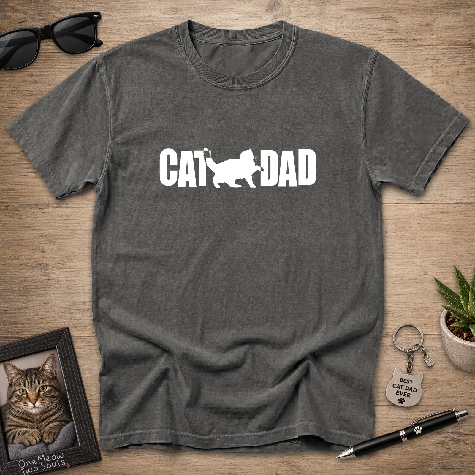 A dark gray t-shirt with a white 'CAT DAD' graphic printed on the chest, laid flat on a wooden surface with lifestyle props including sunglasses, a framed cat photo, a small plant, and a pen.