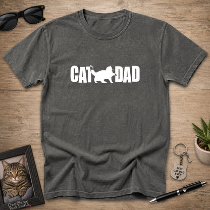 A dark gray t-shirt with a white 'CAT DAD' graphic printed on the chest, laid flat on a wooden surface with lifestyle props including sunglasses, a framed cat photo, a small plant, and a pen.