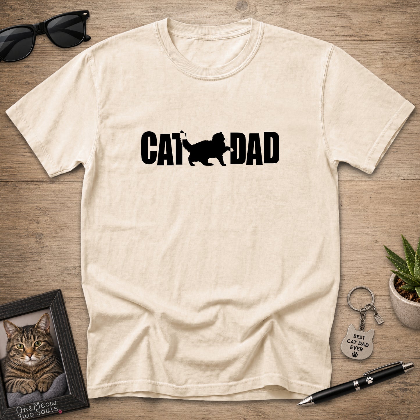 A cream-colored short-sleeve t-shirt with a black graphic reading 'CAT DAD' and a cat silhouette, laid flat on a wooden surface with sunglasses, a framed cat photo, a keychain, and a pen around it.
