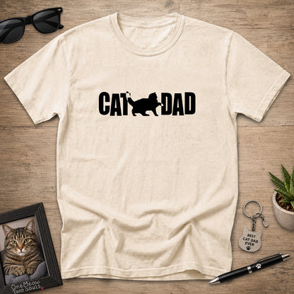 A cream-colored short-sleeve t-shirt with a black graphic reading 'CAT DAD' and a cat silhouette, laid flat on a wooden surface with sunglasses, a framed cat photo, a keychain, and a pen around it.