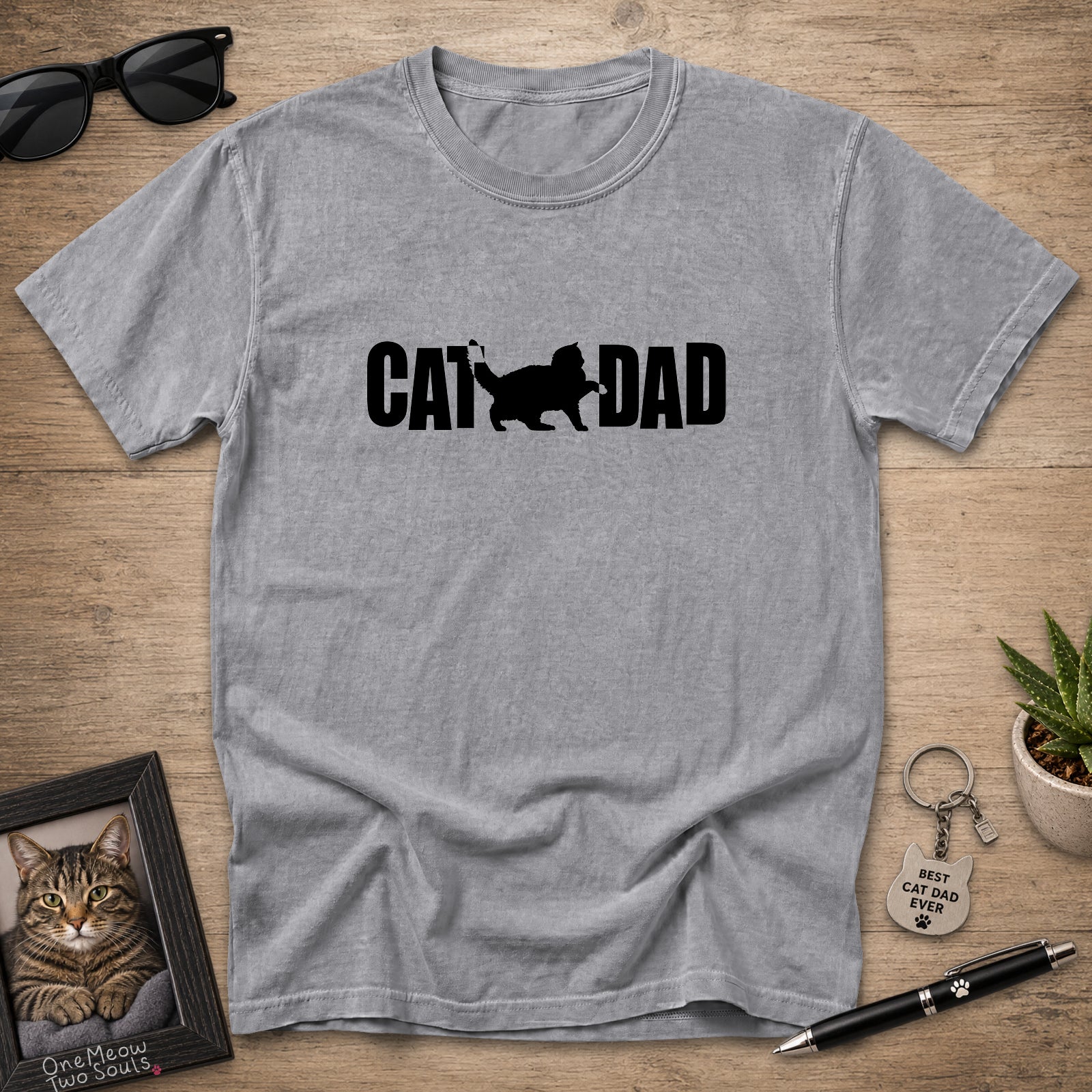 A heather gray short-sleeve t-shirt laid flat on a wooden surface, featuring a black graphic print that reads 'CAT DAD' with a cat silhouette. The shirt is styled with sunglasses, a framed cat portrait, a small potted succulent, a keychain, and a pen.