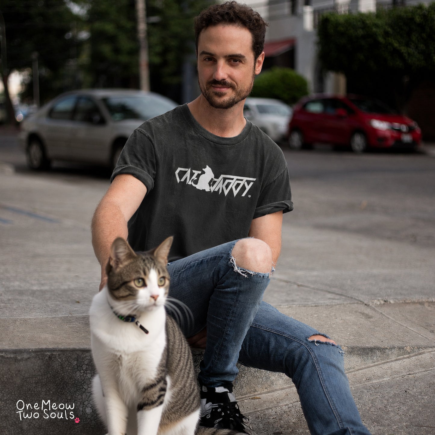 A man sitting on a city sidewalk next to a white and gray tabby cat, wearing a dark gray graphic t-shirt with the text 'Cat Daddy' and ripped blue jeans.
Black Comfort Colors 1717 t-shirt with a "Cat Daddy" graphic. One Meow Two Souls brand.