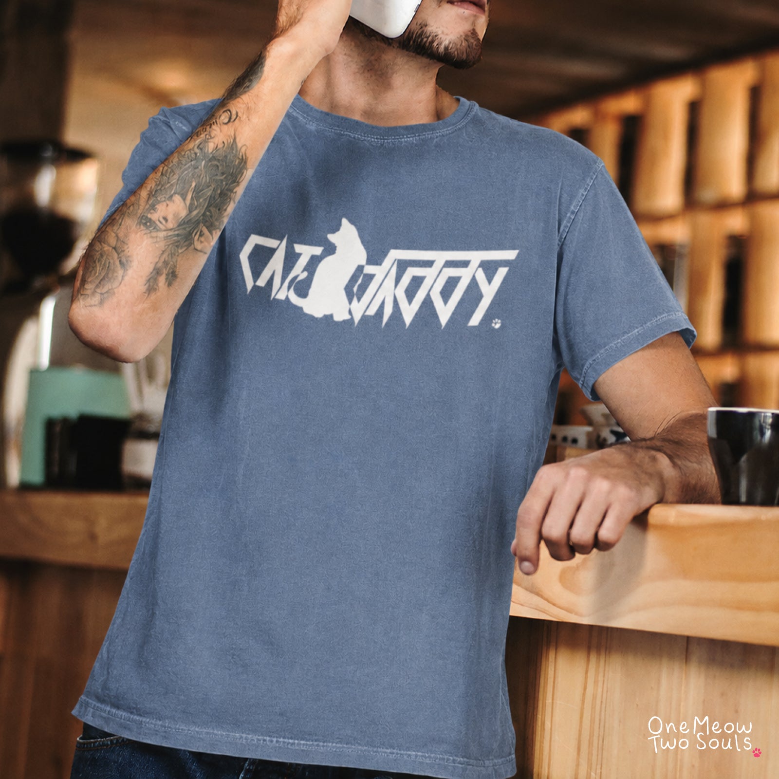 A man wearing a faded blue crewneck t-shirt with a white graphic print that reads 'CAT DADDY' in a stylized font with a cat silhouette. The photo is taken in a warm, wood-toned indoor environment, likely a cafe or studio.
Blue Jean Comfort Colors 1717 t-shirt with a "Cat Daddy" graphic. One Meow Two Souls brand.