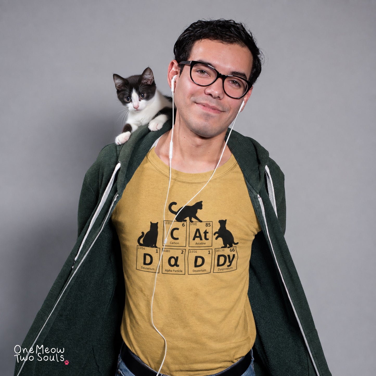 A man wearing glasses and a dark green zip-up hoodie over a mustard yellow graphic t-shirt with a cat-themed 'Cat daddy' design, with a black and white cat sitting on his shoulder against a gray studio background.
Mustard color Comfort Colors 1717 t-shirt tee with a Cat Daddy chemisty periodic table design. One Meow Two Souls brand.