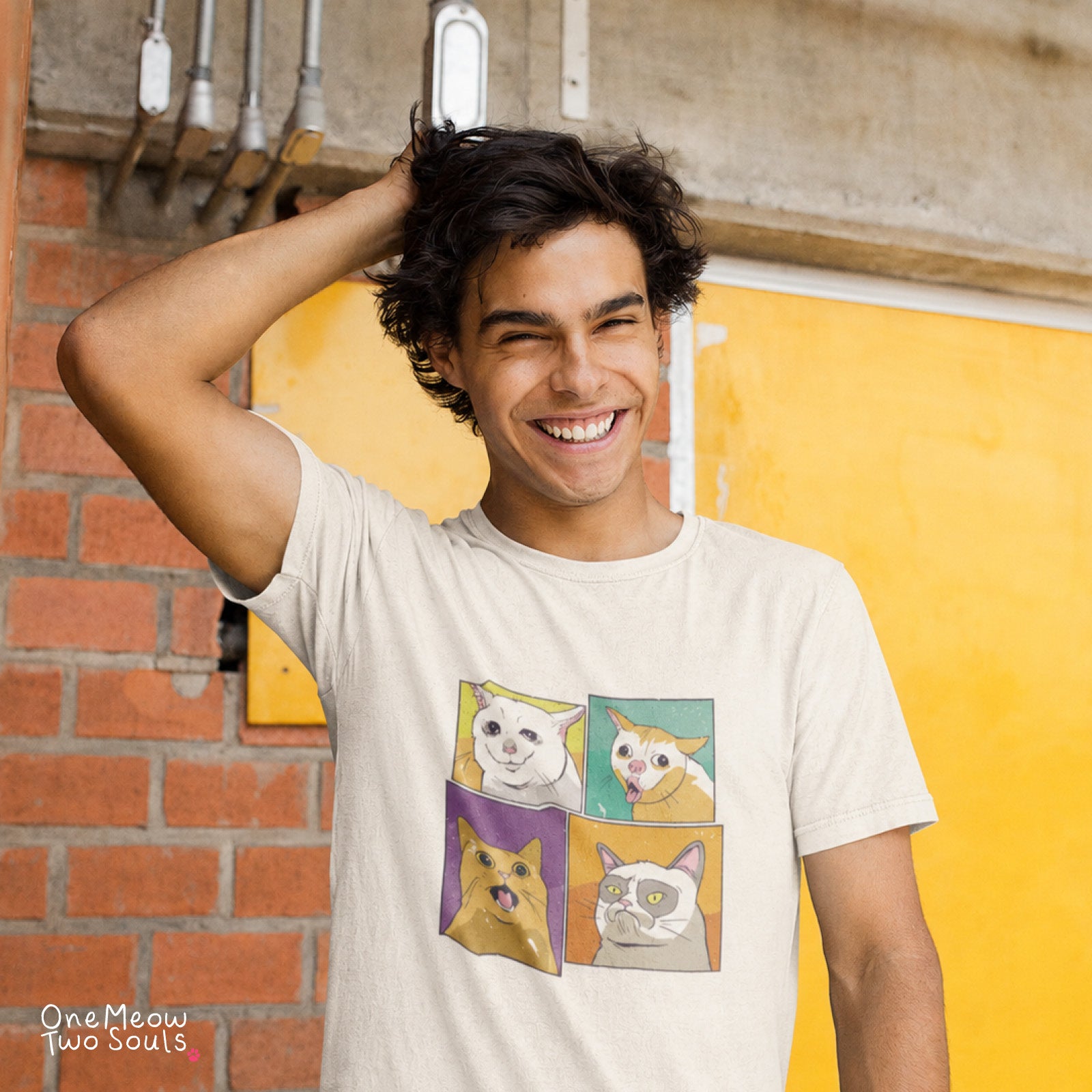 A smiling young man wearing a light beige graphic t-shirt with a four-panel cat illustration, standing in front of a yellow door and brick wall. The watermark 'One Meow Two Souls' is visible in the bottom-left corner.
Ivory colored Comfort Colors 1717 cotton T-short featuring a colorful Cat Meme faces design. One Meow Two Souls brand.