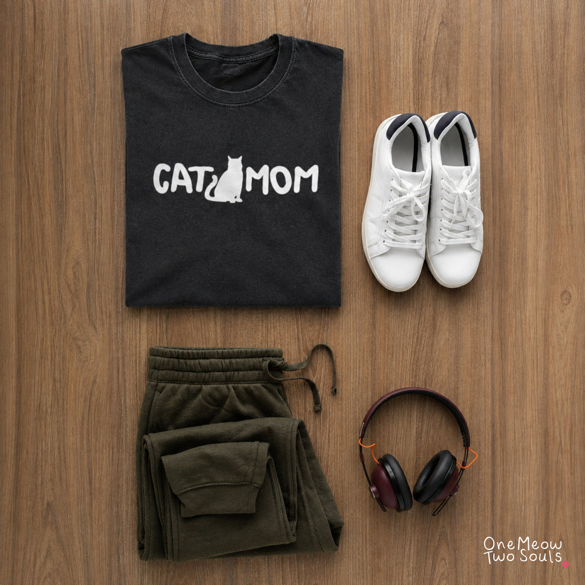 A styled flat-lay of a black t-shirt with a white 'CAT MOM' graphic, paired with olive green sweatpants, white sneakers, and headphones on a wooden background. The brand 'One Meow Two Souls' is visible in the bottom right corner.