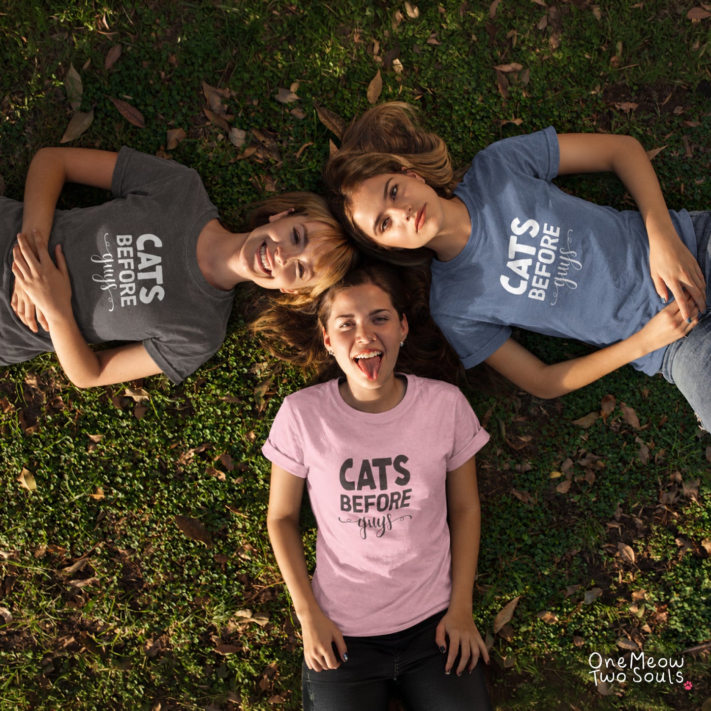 Three women lying on grass in a circle, each wearing a graphic T-shirt with the text 'CATS BEFORE guy'. The shirts are in different colors: dark gray, medium blue, and light pink. One Meow Two Souls brand.