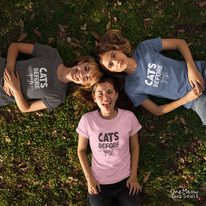 Three women lying on grass in a circle, each wearing a graphic T-shirt with the text 'CATS BEFORE guy'. The shirts are in different colors: dark gray, medium blue, and light pink. One Meow Two Souls brand.