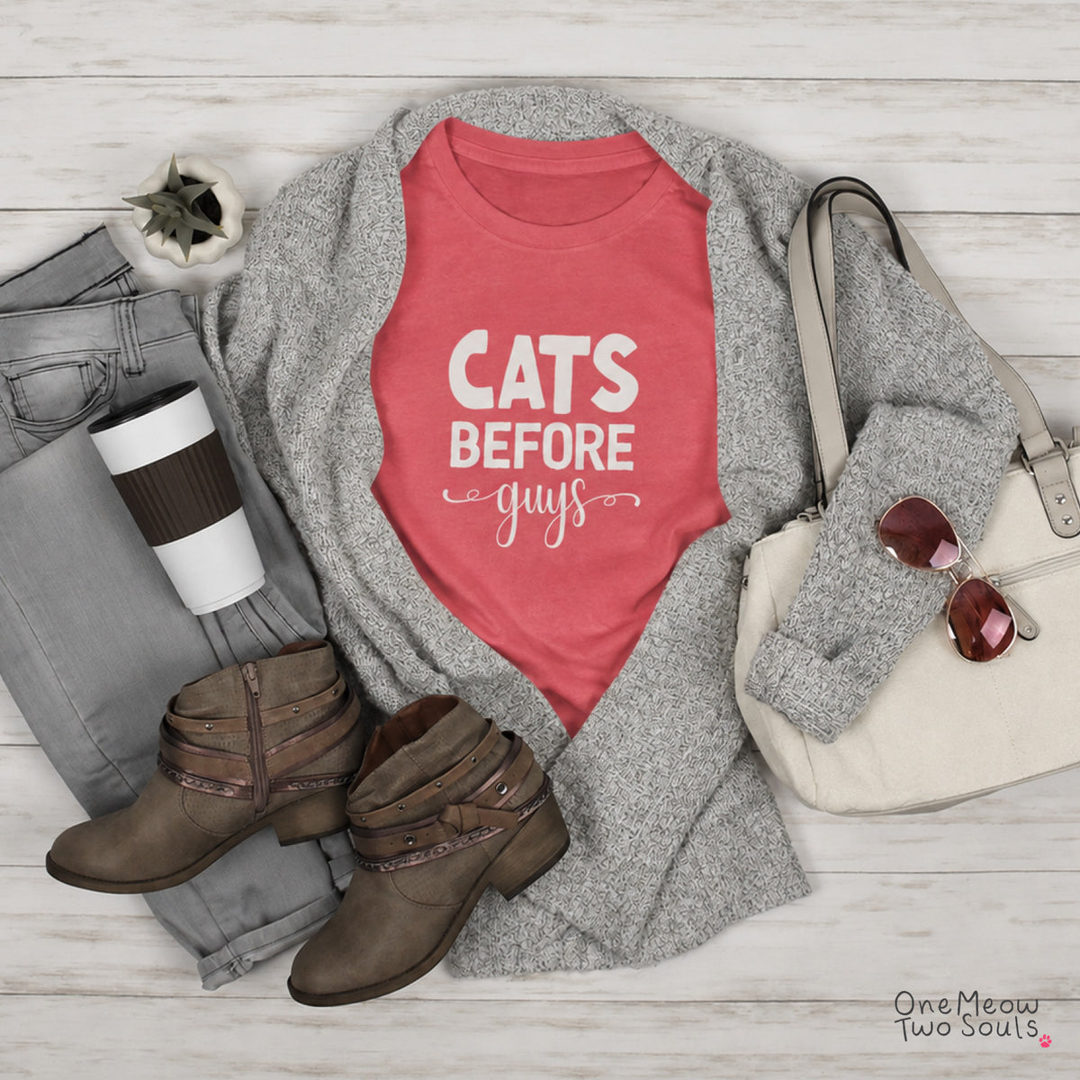 A styled flat lay featuring a coral pink graphic T-shirt with the text 'CATS BEFORE guys' in white, layered under a heather gray knit cardigan. The outfit is arranged on a white wood plank background alongside gray jeans, brown ankle boots, a cream handbag with sunglasses, and a coffee cup. One Meow Two Souls brand.