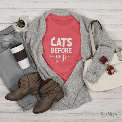 A styled flat lay featuring a coral pink graphic T-shirt with the text 'CATS BEFORE guys' in white, layered under a heather gray knit cardigan. The outfit is arranged on a white wood plank background alongside gray jeans, brown ankle boots, a cream handbag with sunglasses, and a coffee cup. One Meow Two Souls brand.