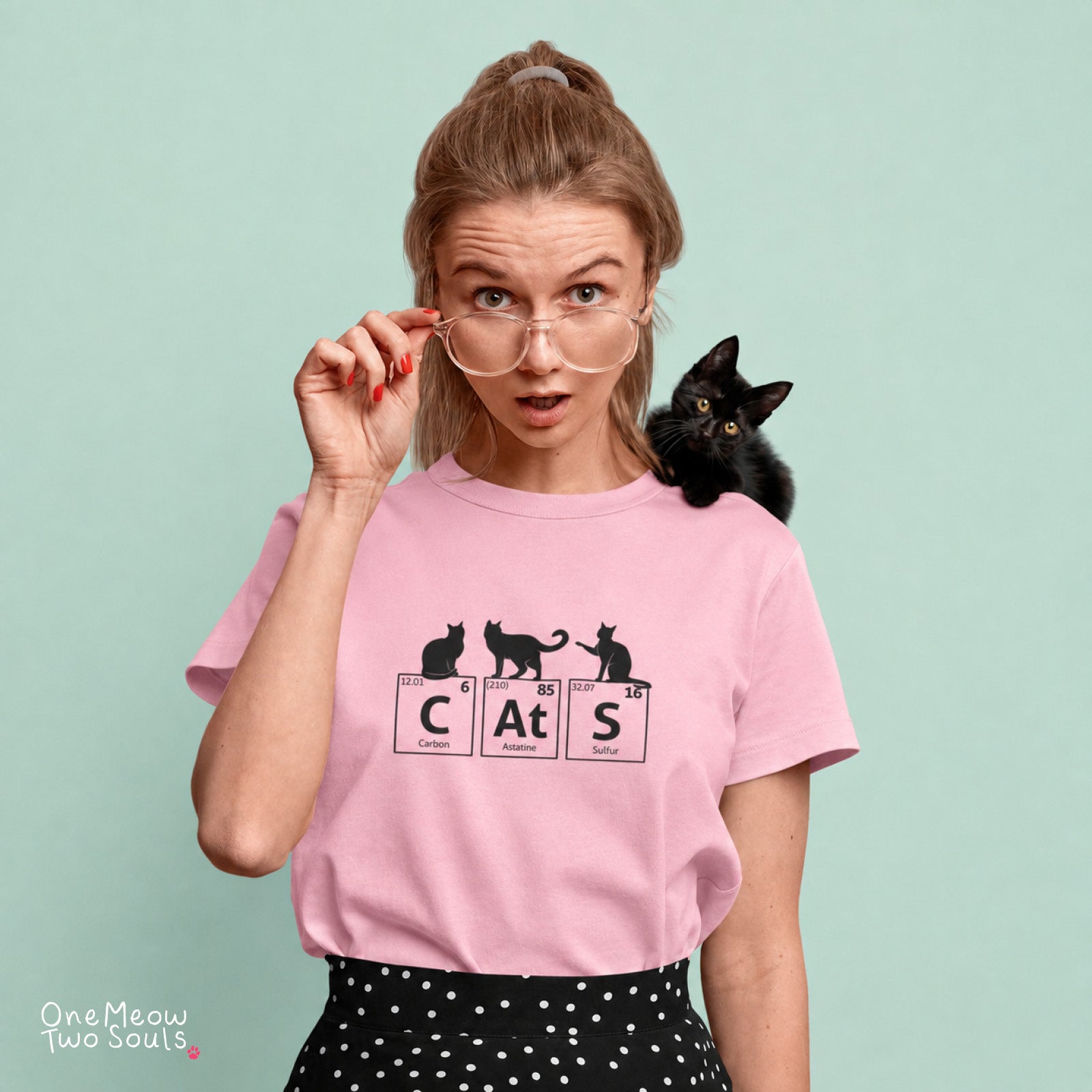 A woman wearing a light pink graphic t-shirt with a black cat-themed print ('CATS' in periodic table style) and a small black kitten on her shoulder, set against a solid mint green background.
Blossom colored Comfort Colors 1717 cotton T-shirt, presenting a "CatS" funny chemistry periodic table cat design. One Meow Two Souls brand.