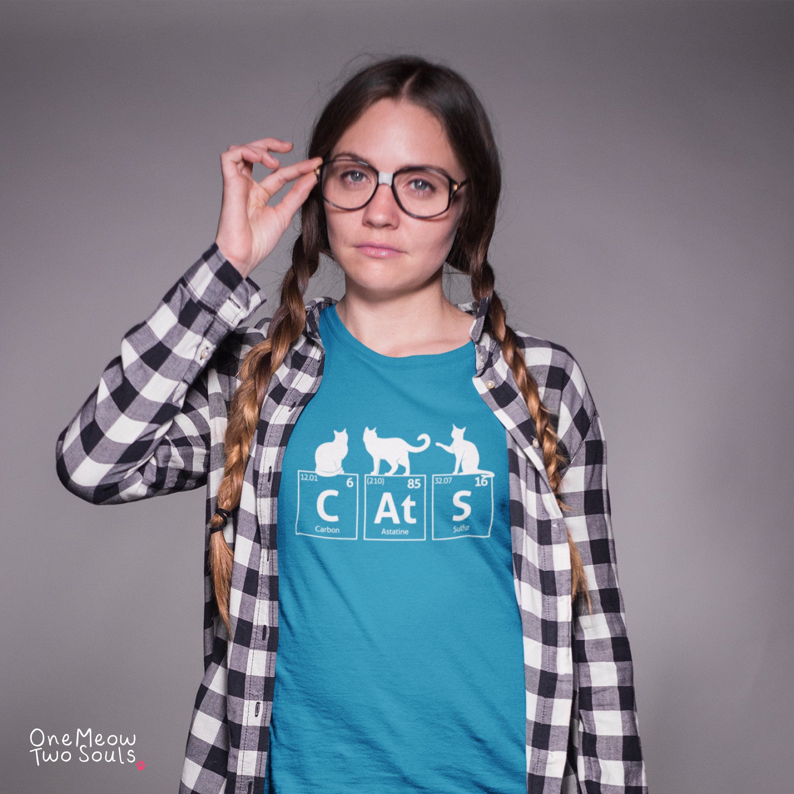 A young woman with braided hair and glasses wears a teal graphic t-shirt with a white cat-themed periodic table design, layered under a black and white plaid flannel shirt, against a solid gray studio background.
Sapphire colored Comfort Colors 1717 cotton T-shirt, presenting a "CatS" funny chemistry periodic table cat design. One Meow Two Souls brand.