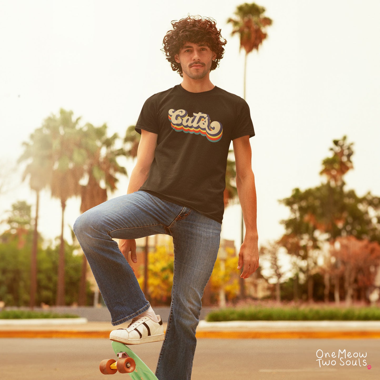 A man with curly hair stands on a skateboard in an outdoor park setting, wearing a black graphic t-shirt with a retro rainbow 'Cats' design, blue jeans, and white sneakers. The background features warm, golden-hour lighting and blurred palm trees.
Black colored Comfort Colors 1717 t-shirt with a retro "Cats" typography design. Onw Meow Two Sould brand.