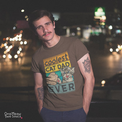 A man wearing a brown graphic t-shirt with a 'Coolest Cat Dad' print, standing outdoors at night with blurred city lights in the background. The brand 'One Meaw Two Souls' is visible in the bottom-left corner.